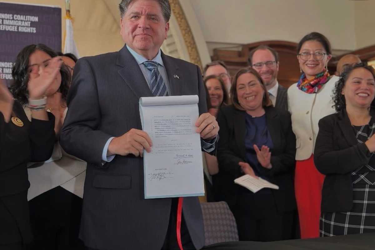 Governor JB Pritzker holding the signed HB 1312 bill while standing with advocates, decisionmakers, and community members applauding during the bill signing event.
