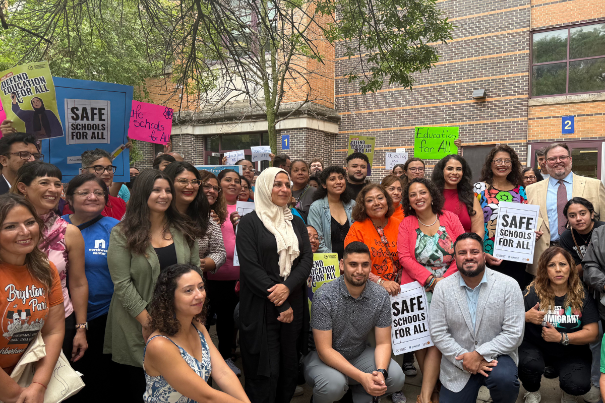 Group of people at an outdoor rally holding signs with messages supporting education, safety, and immigrant rights.