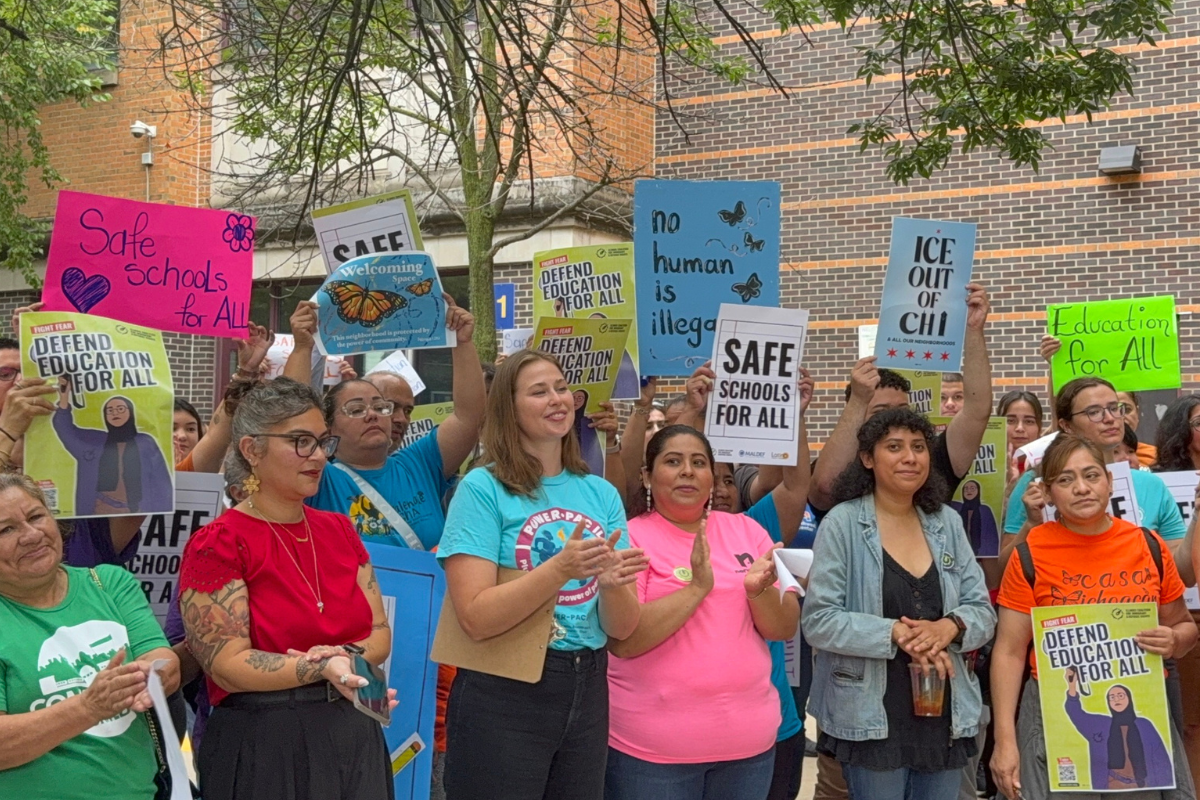 A group of people at a rally holding signs that read “Defend Education for All,” “Safe Schools for All,” and “No Human is Illegal.”
