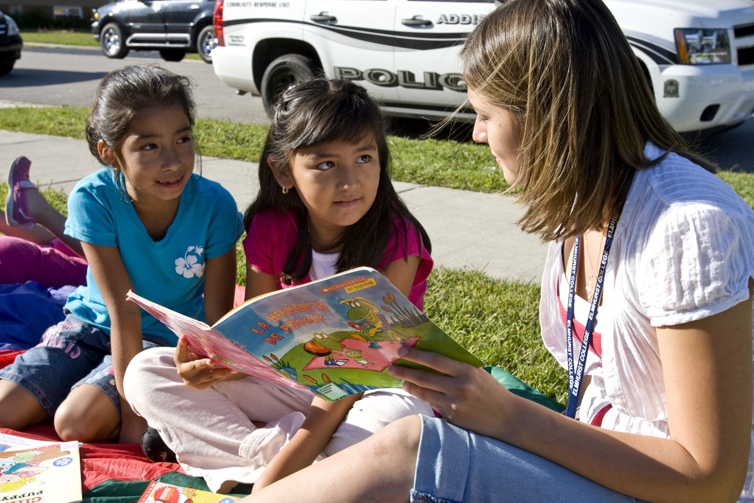 teacher-reading-outdoors-with-students A teacher reads a Spanish children’s book to two young students sitting on a blanket outside.